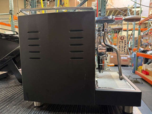 Close view of a LUCCA M58 espresso machine with black body chrome group head and wooden portafilter handles on a warehouse shelf