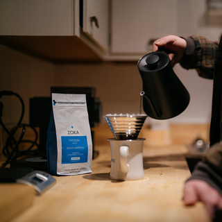 Person pours hot water over a pour over setup with a Zoka Tatoosh bag visible in the background