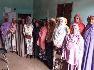 Group of women wearing colorful hijabs and robes standing together in a green walled room.