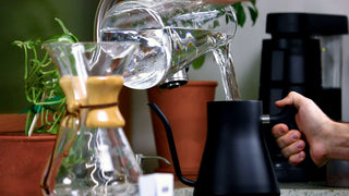 Hand pours hot water from a gooseneck kettle into a glass coffee server on a kitchen counter